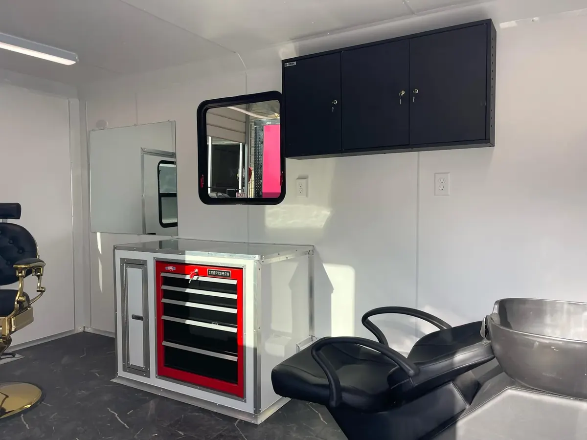 Barbershop trailer interior with a black shampoo chair and bowl, a red Craftsman tool chest cabinet, black wall cabinets, a wall mirror and the edge of a barber chair