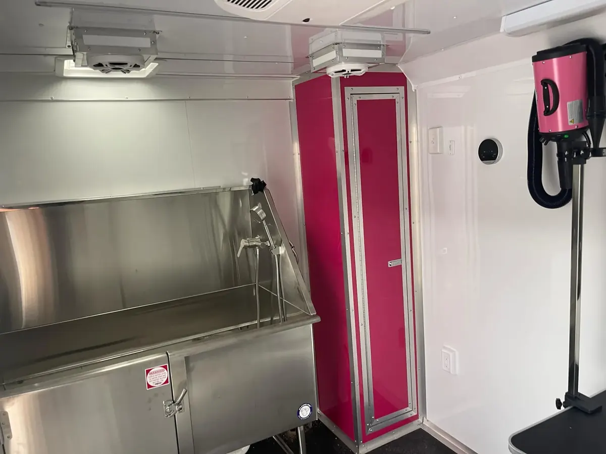 Dog grooming trailer interior with a stainless steel tub, a tall pink cabinet and a pink dryer mounted beside the grooming table