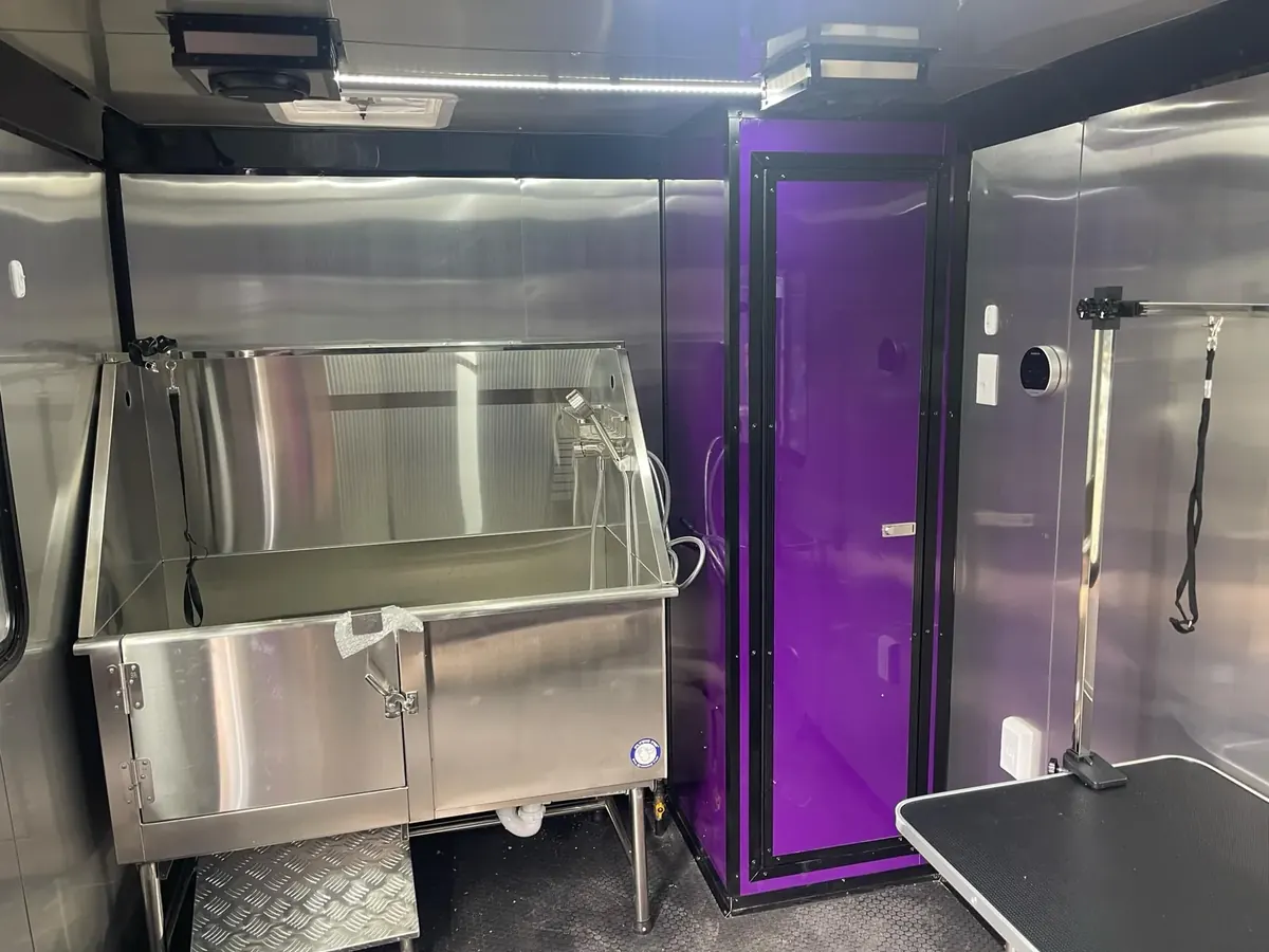 Dog grooming trailer interior with stainless steel walls, a stainless steel tub with a diamond-plate step, a tall purple cabinet and a grooming table with a grooming arm